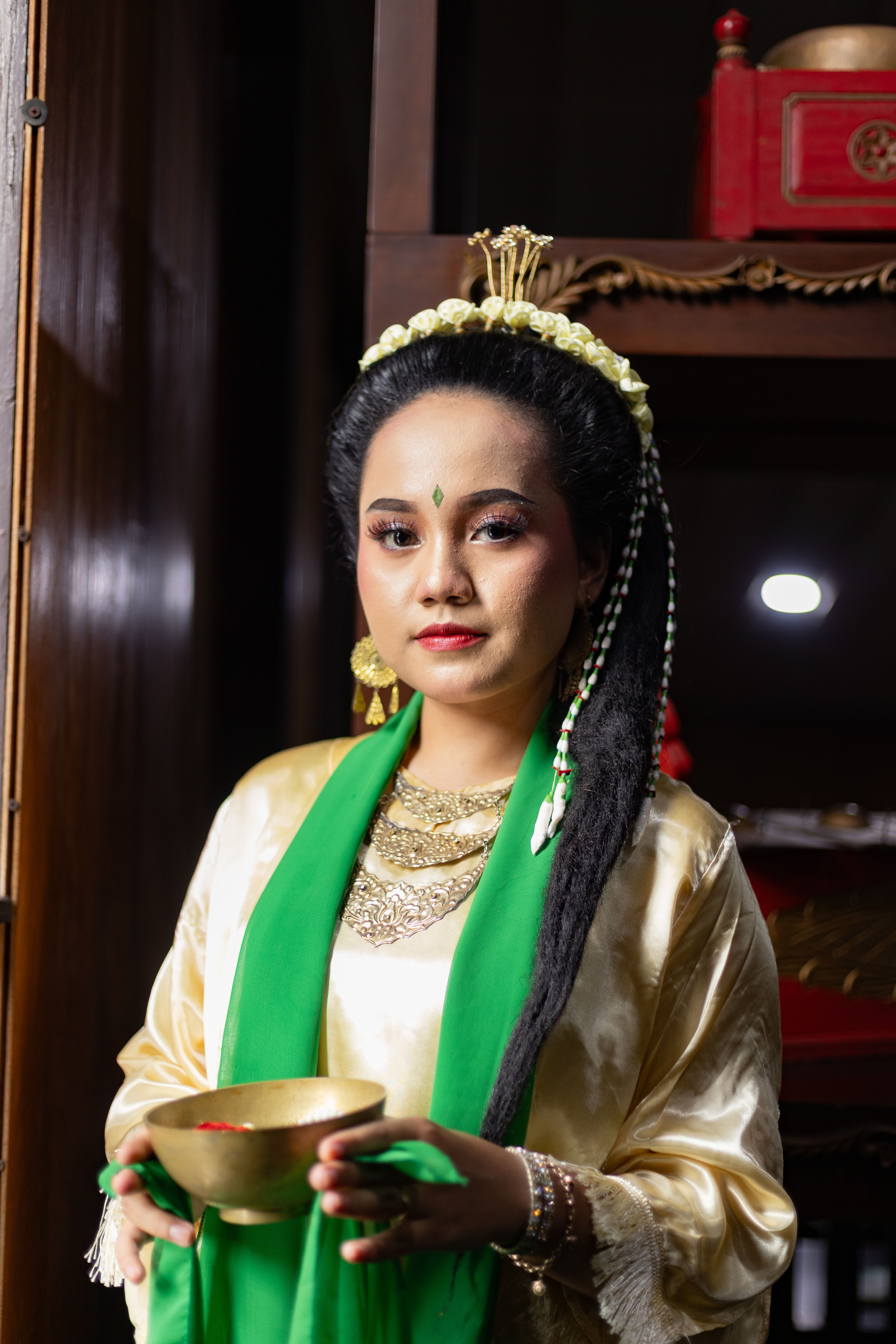 A woman in traditional dress holding a bowl, adorned with gold jewelry and elaborate hairstyle.