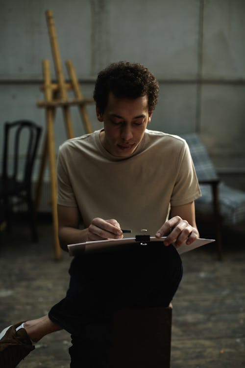 A Man Sitting On Wooden Chair Sketching On White Cardboard