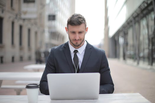 Professional man in a suit using a laptop and earphones while sitting outdoors.