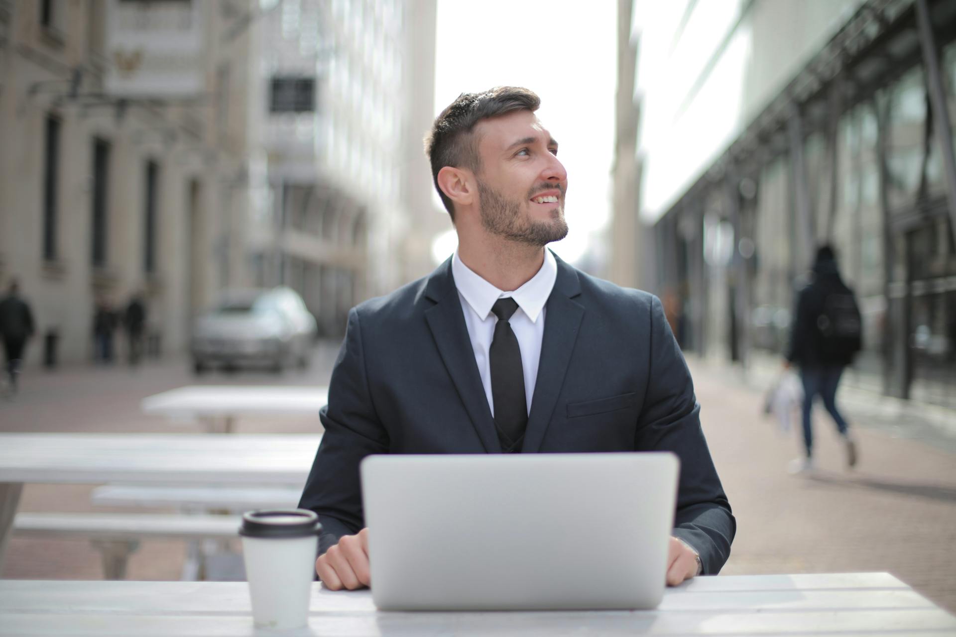 Smiling businessman in formal attire working on a laptop outdoors, exuding confidence and happiness.