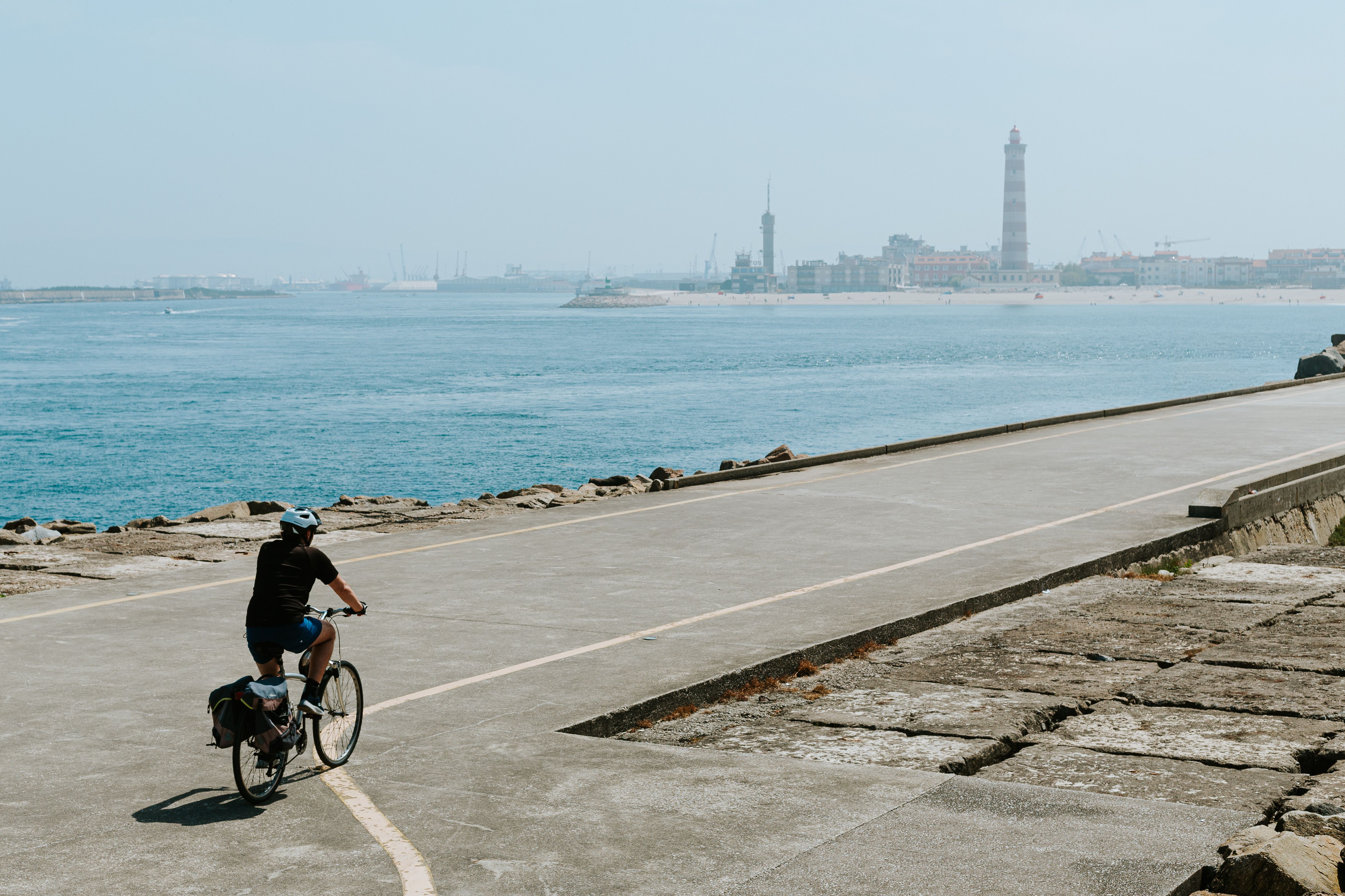 Person cycling along a coastal path with lighthouse and cityscape in view.