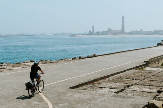 Person cycling along a coastal path with lighthouse and cityscape in view.