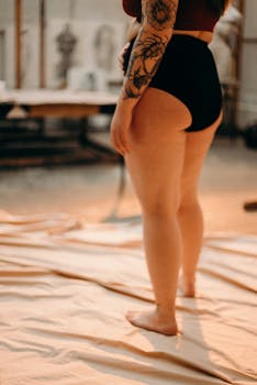 Artistic nude woman standing in a studio environment, showcasing body tattoos and confidence.