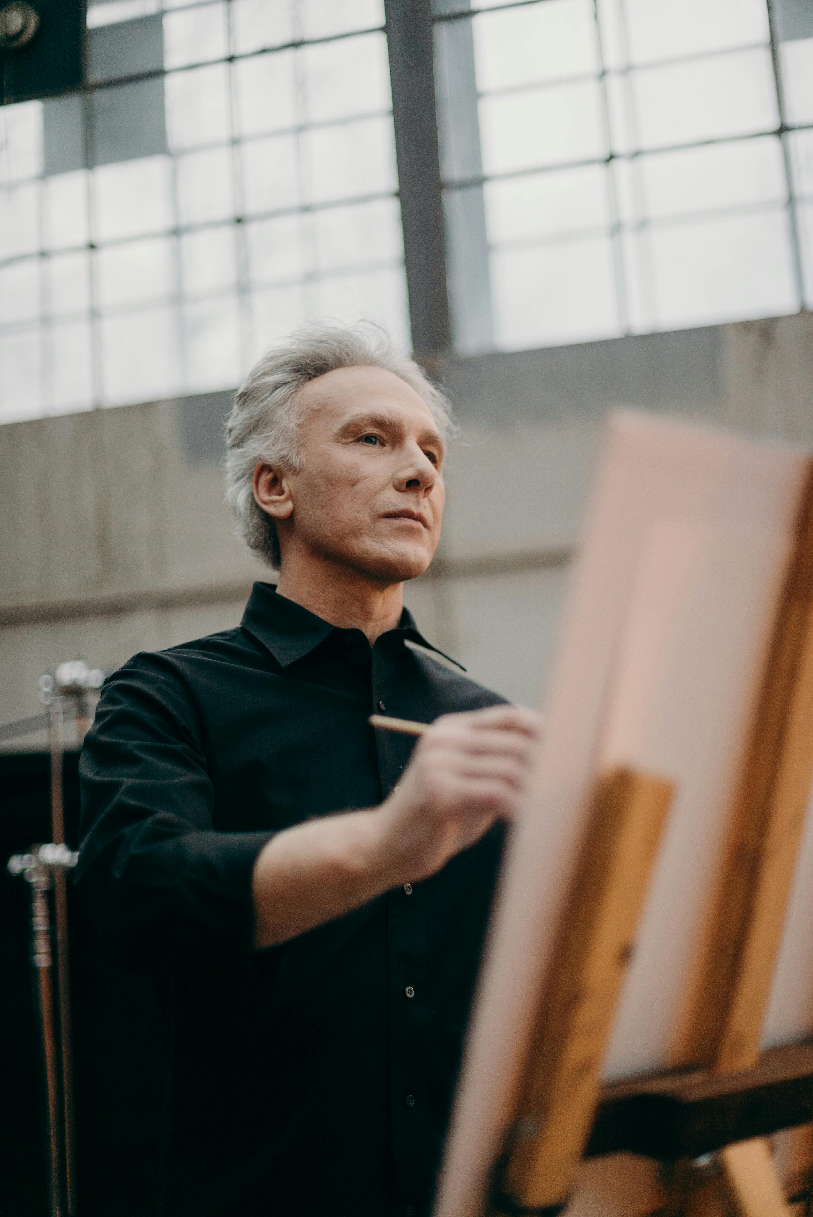 一位身穿黑色衬衫的老人，在光线充足的室内，若有所思地在画架上素描