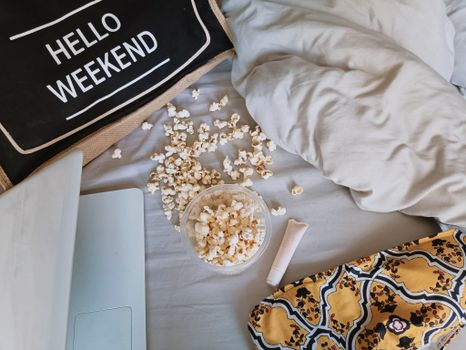 Relaxing weekend scene with popcorn, a laptop, and a 'Hello Weekend' pillow on a bed.