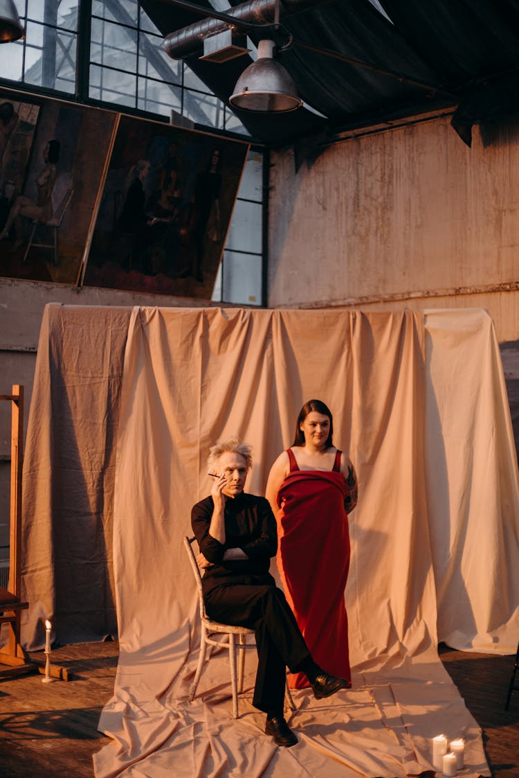 Woman In Red Dress Standing Beside On Man In Black Long Sleeve Seated On A Chair