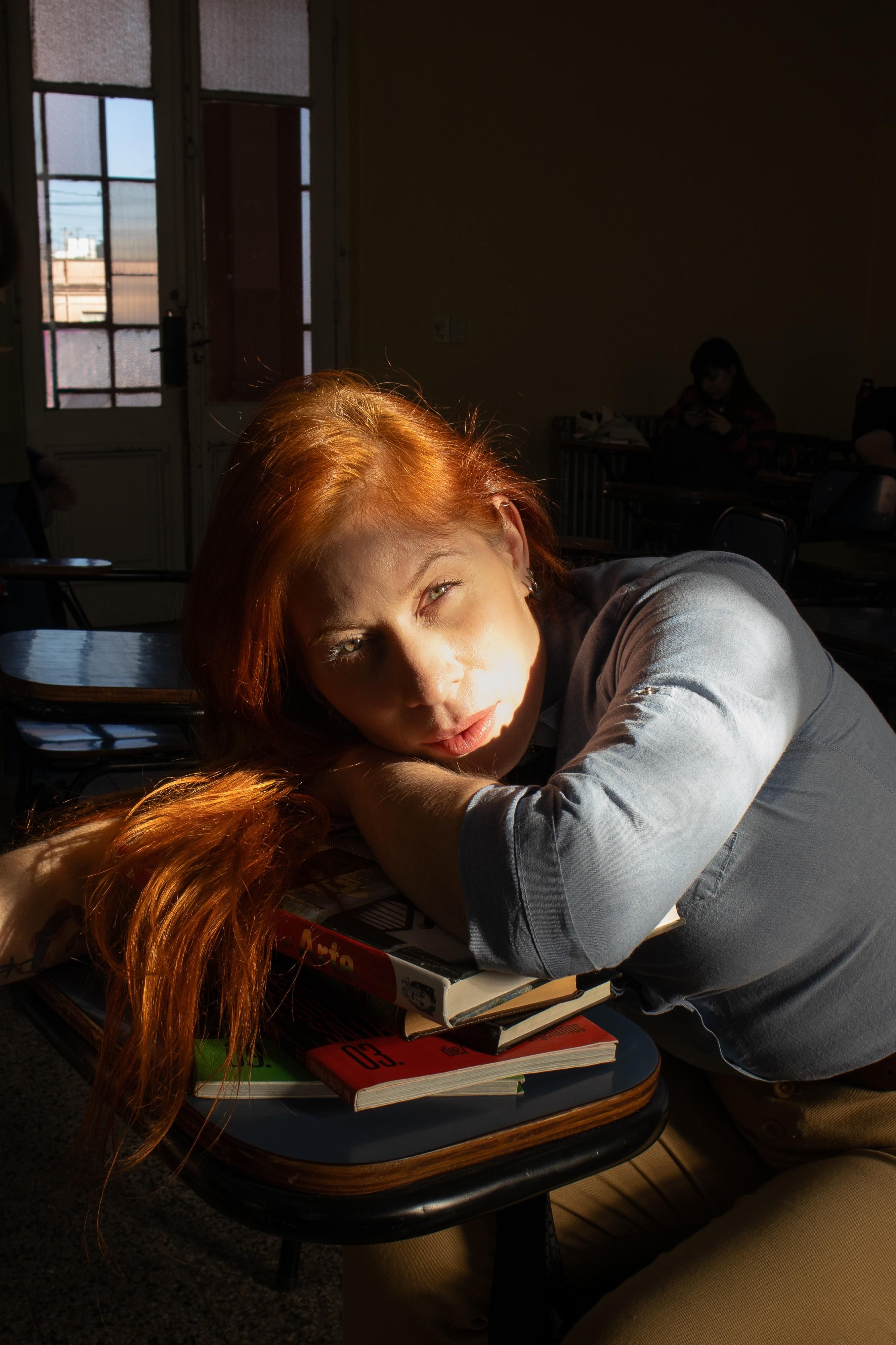Woman resting on books in a sunlit classroom, evoking introspection.