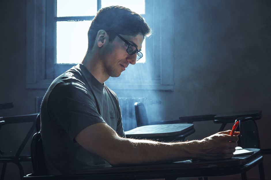 A young adult focused on writing in a dimly lit classroom with dramatic window lighting. A young adult focused on writing in a dimly lit classroom with dramatic window lighting.