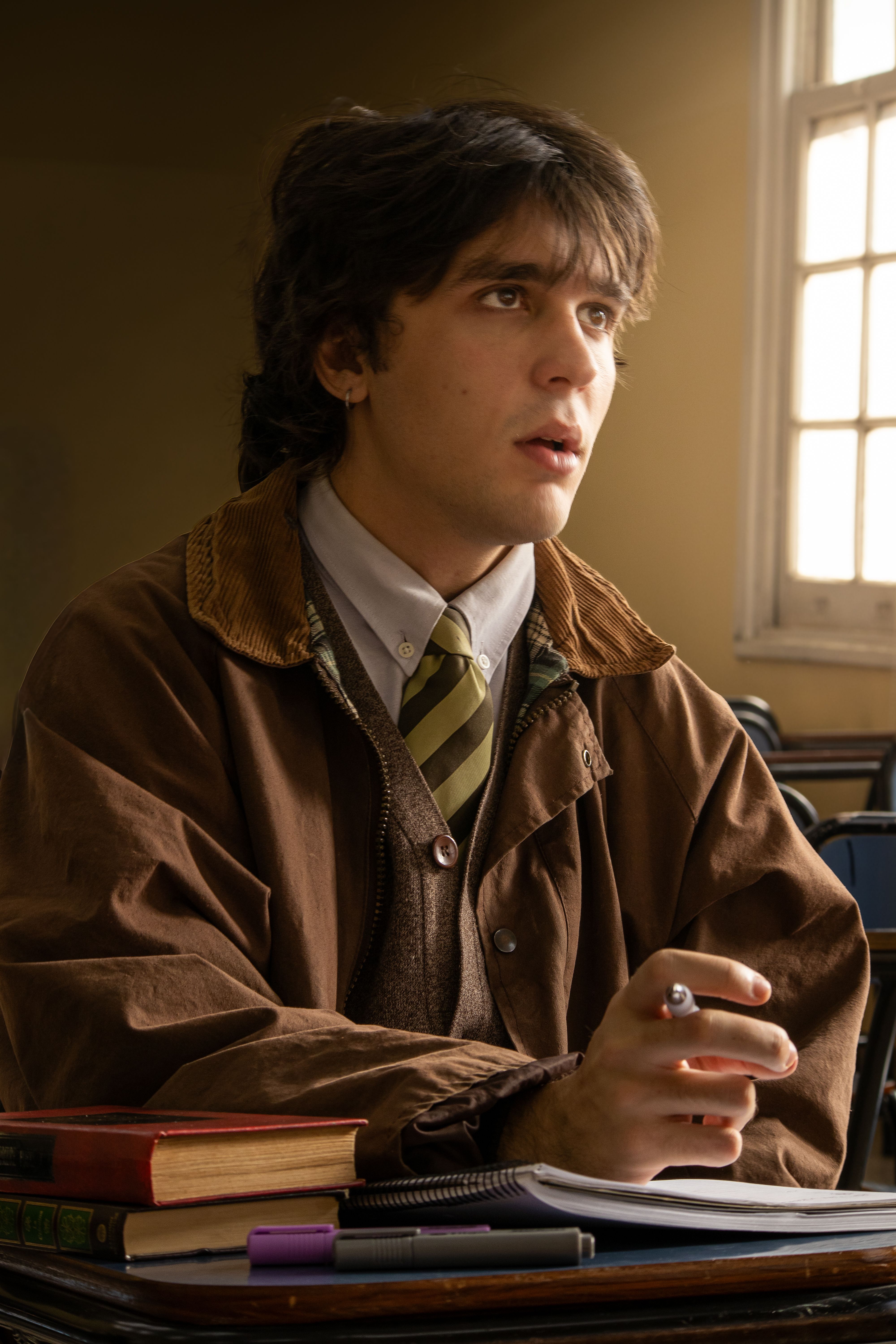 A student in a classroom, deep in thought with books and stationery.