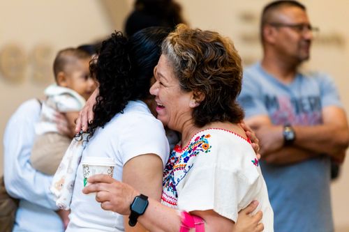 Free Two women embrace joyfully in a bright room, surrounded by family. Warmth and happiness fill the scene. Stock Photo