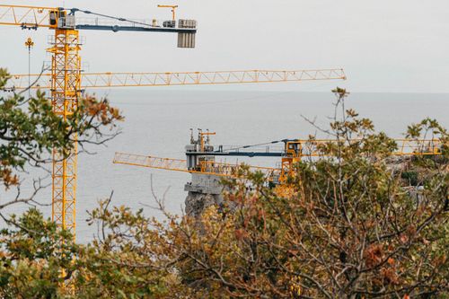 Free Construction site with tower cranes by the ocean, surrounded by autumn foliage. Stock Photo