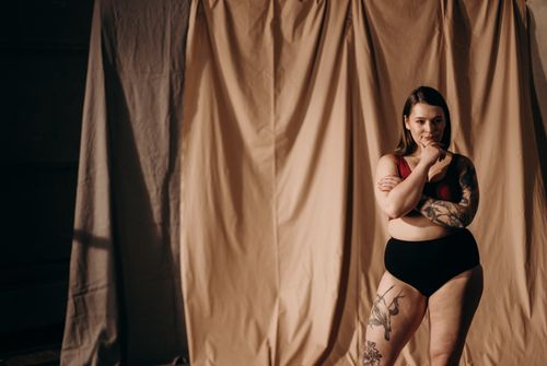 Free Fashion portrait of a confident woman posing in underwear with tattoos, set against a draped fabric backdrop. Stock Photo