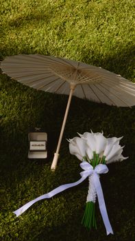 A serene wedding scene with a parasol, bouquet, and rings on grass.