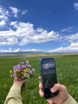 Hands holding a phone and wildflowers against a vast green field and blue sky.