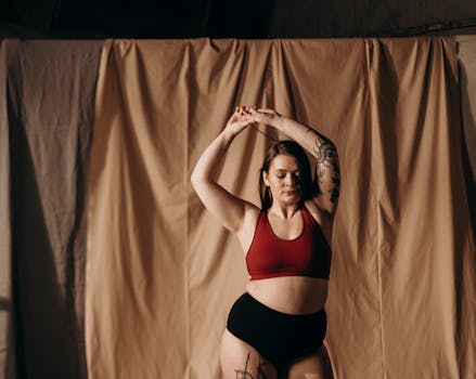 A tattooed woman poses gracefully with closed eyes, standing against a neutral backdrop.