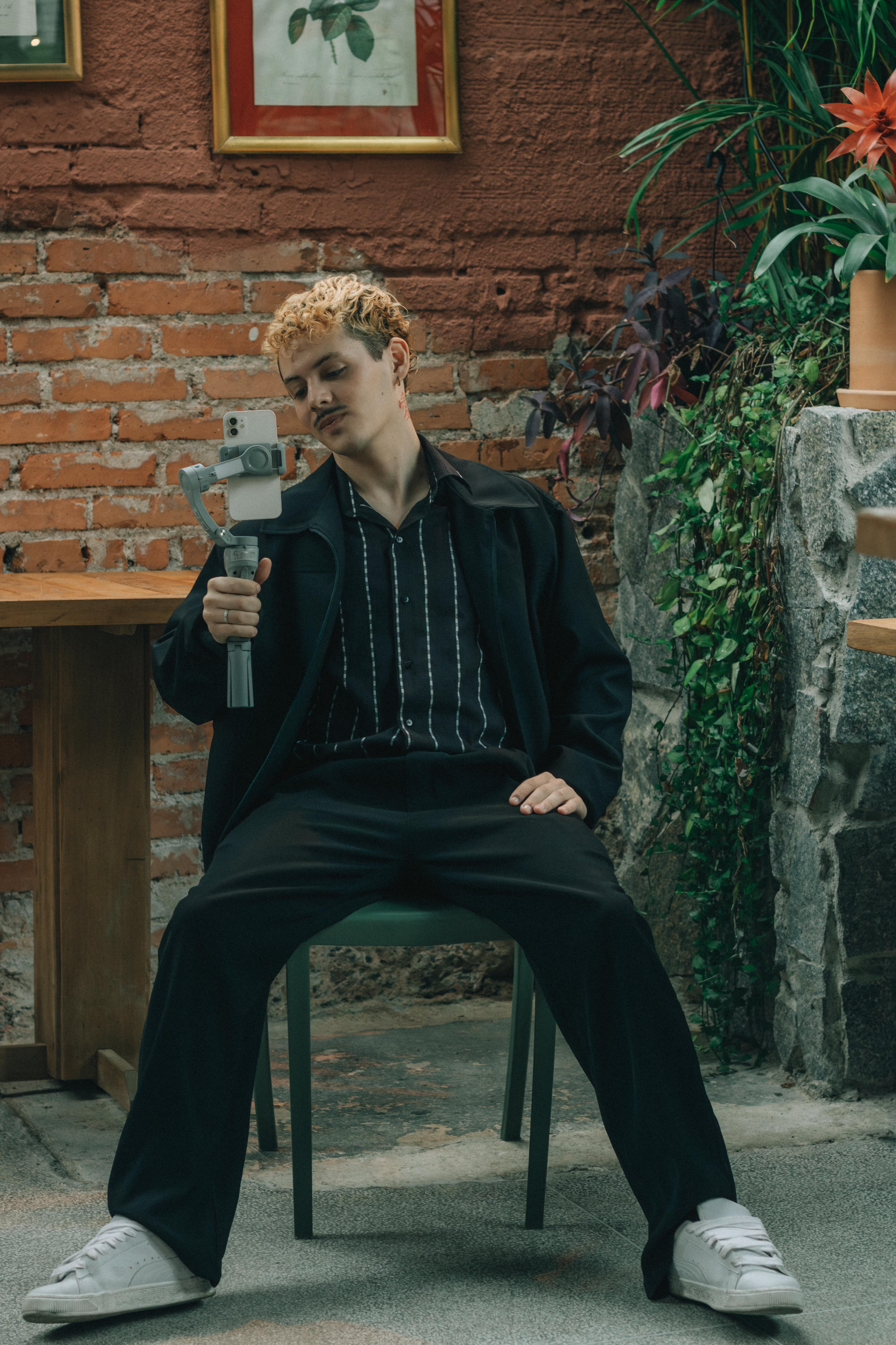Young adult sitting indoors with camera amidst cozy decor and brick backdrop.
