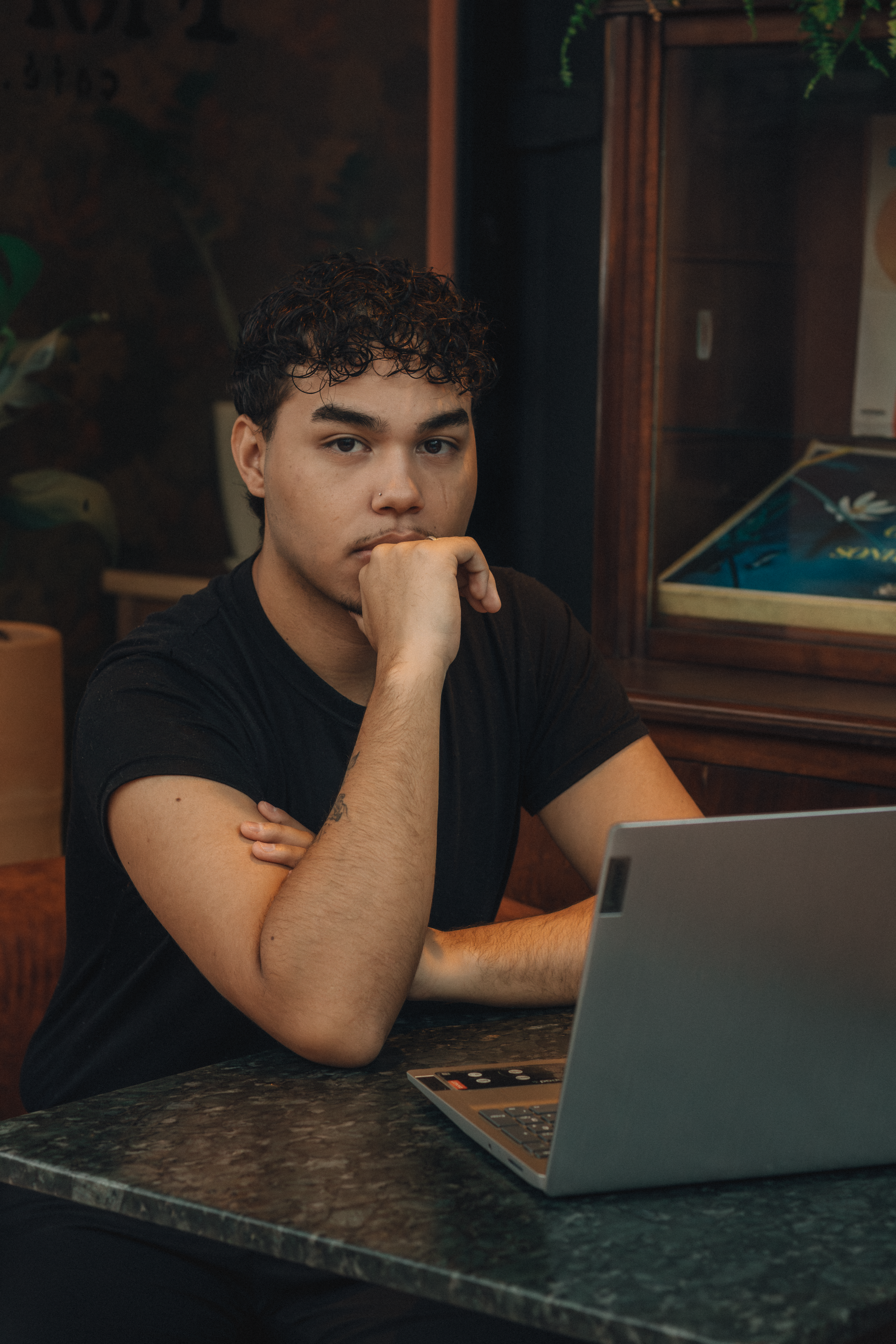 A young man sits thoughtfully at a laptop in a warm, cozy café setting, conveying concentration.