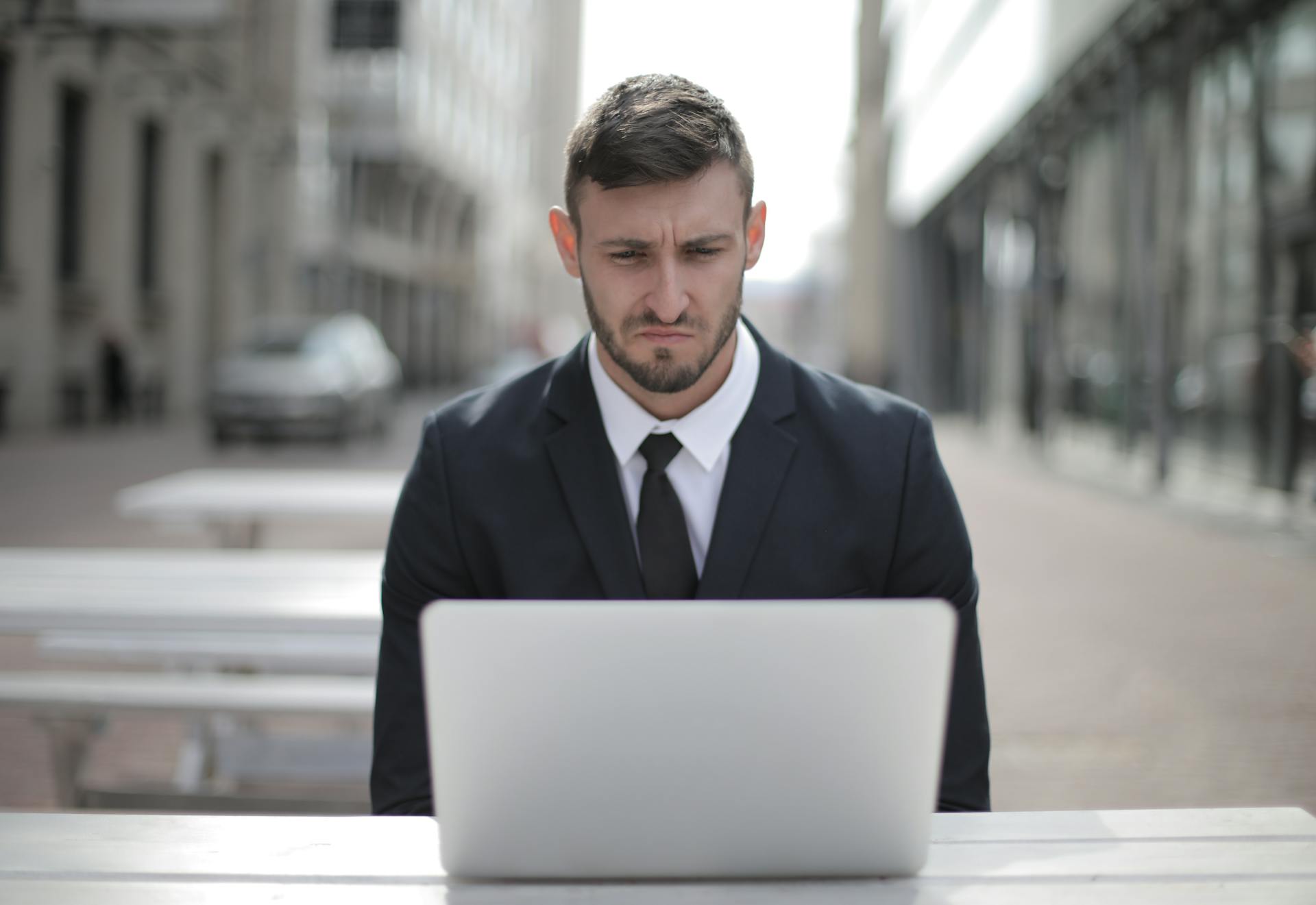 A focused businessman in a suit works on his laptop outdoors in an urban setting.