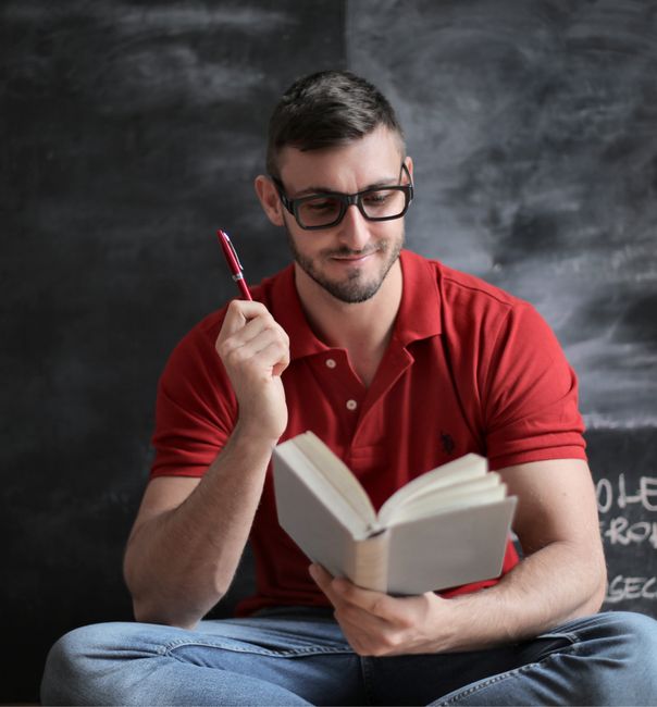 Young man in glasses reading a book attentively, holding a pen. Young man in glasses reading a book attentively, holding a pen.