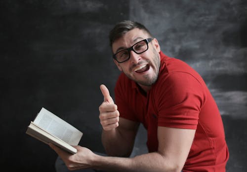 Free Man in Red Crew Neck T-shirt Wearing Black Framed Eyeglasses Holding White Book Stock Photo