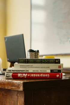 Stack of books on a wooden desk with a laptop in a classroom setting, Buenos Aires.