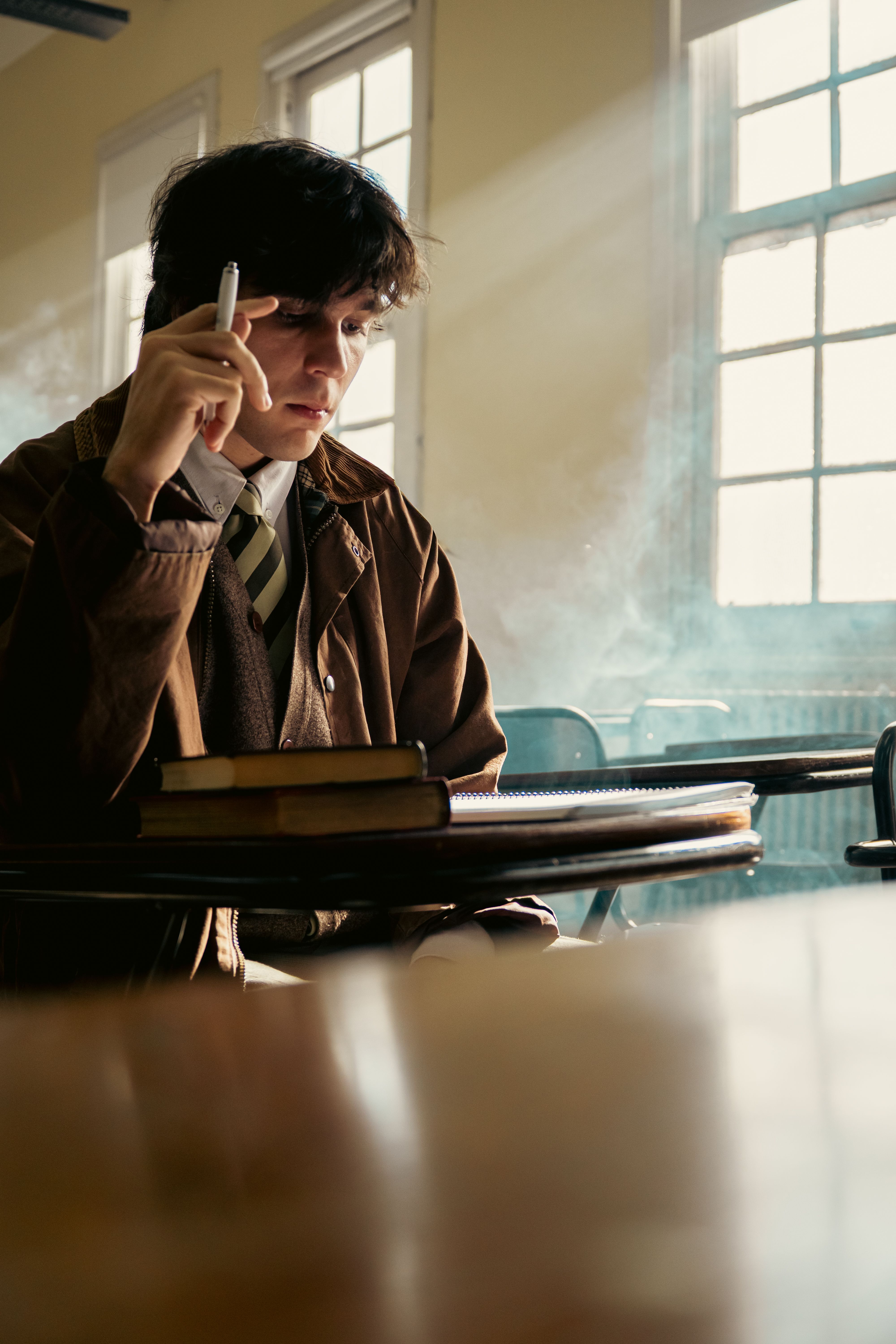 Young man in a classroom with books and natural light, pondering.
