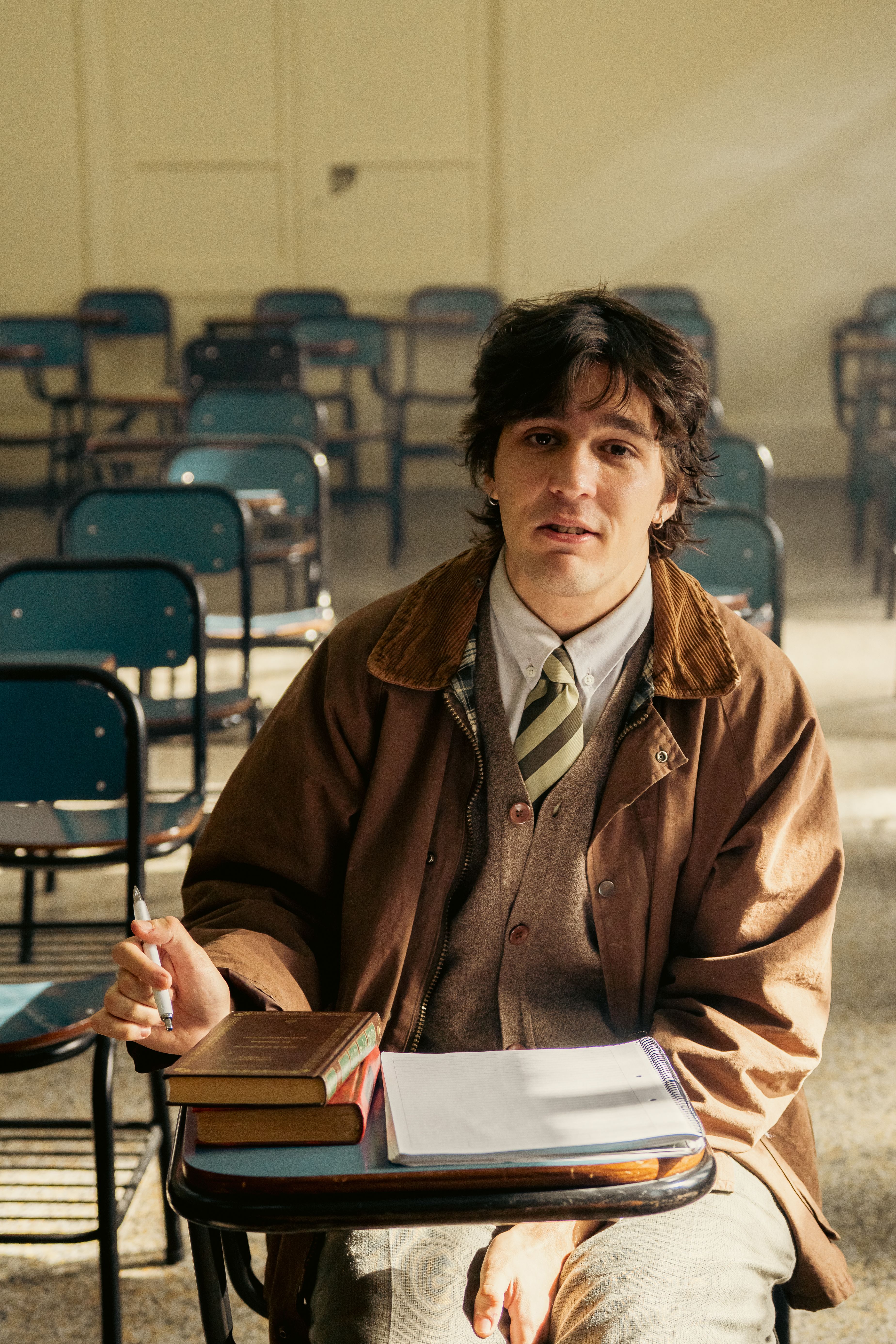 A university student sitting in a Buenos Aires classroom, holding a pen and with books on the desk.