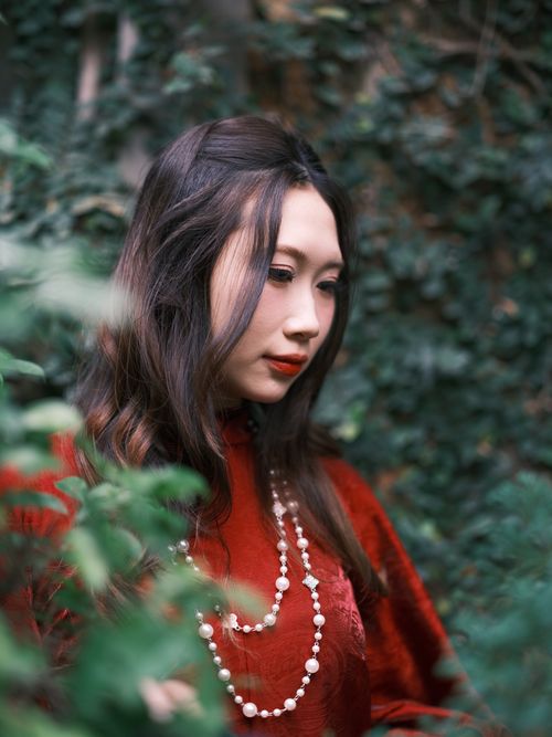 Free Elegant portrait of a woman in a red dress and pearl necklace, surrounded by lush greenery. Stock Photo