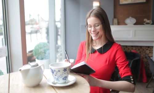 Free Smiling woman in red dress enjoying tea and writing in a cafe by the window. Stock Photo