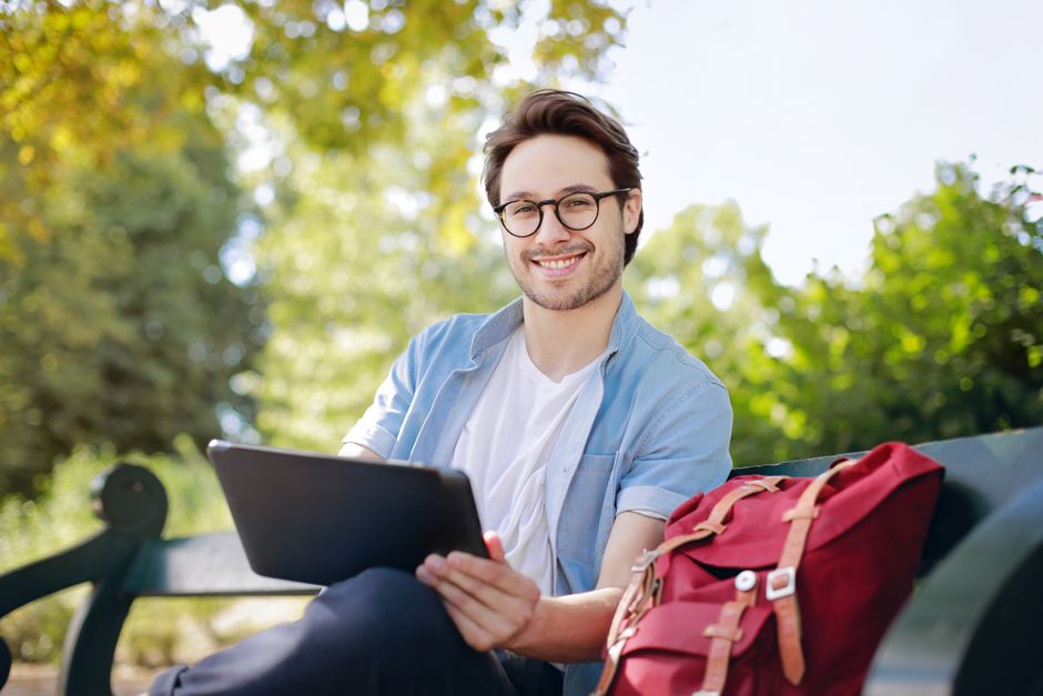 Smiling young man with glasses using a tablet on a park bench with a red backpack nearby. Smiling young man with glasses using a tablet on a park bench with a red backpack nearby.
