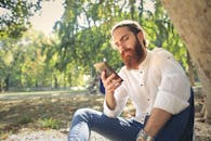 一名留着胡须、穿着白衬衫的男子坐在公园外使用智能手机