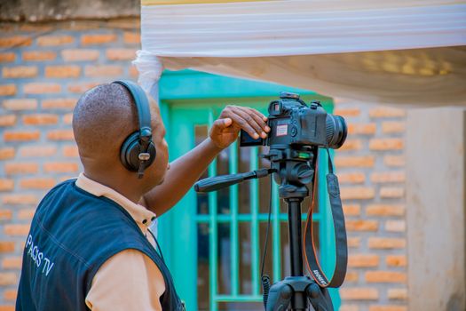 Cameraman wearing headphones operates DSLR on tripod indoors.