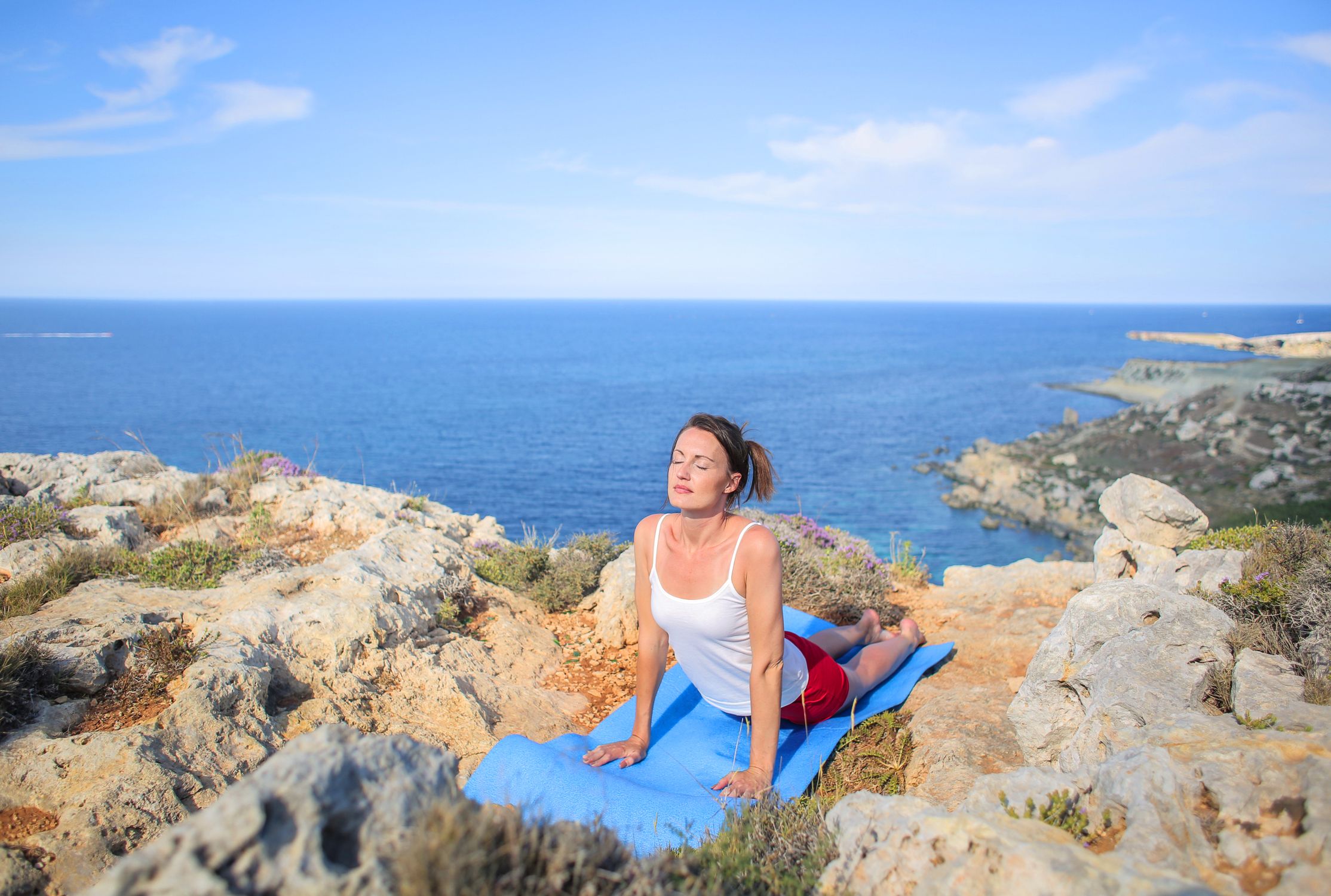 Woman in white tank top practicing yoga on a rocky shore with ocean backdrop