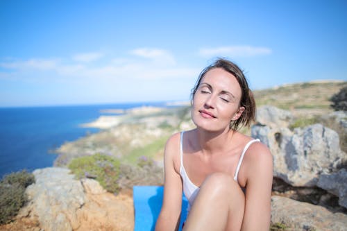 Free Woman in White Tank Top Sitting on Brown Rock Under the Blue Sky Stock Photo