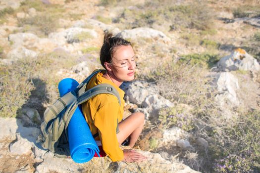 Relaxed woman with backpack and yoga mat sits on rocky terrain during summer hike.