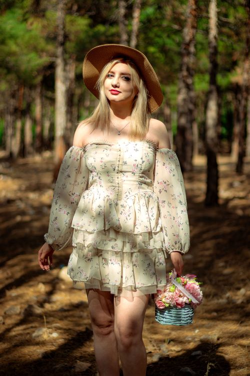Free Blonde woman in a floral dress and hat carrying a basket while walking in a sunlit forest. Stock Photo