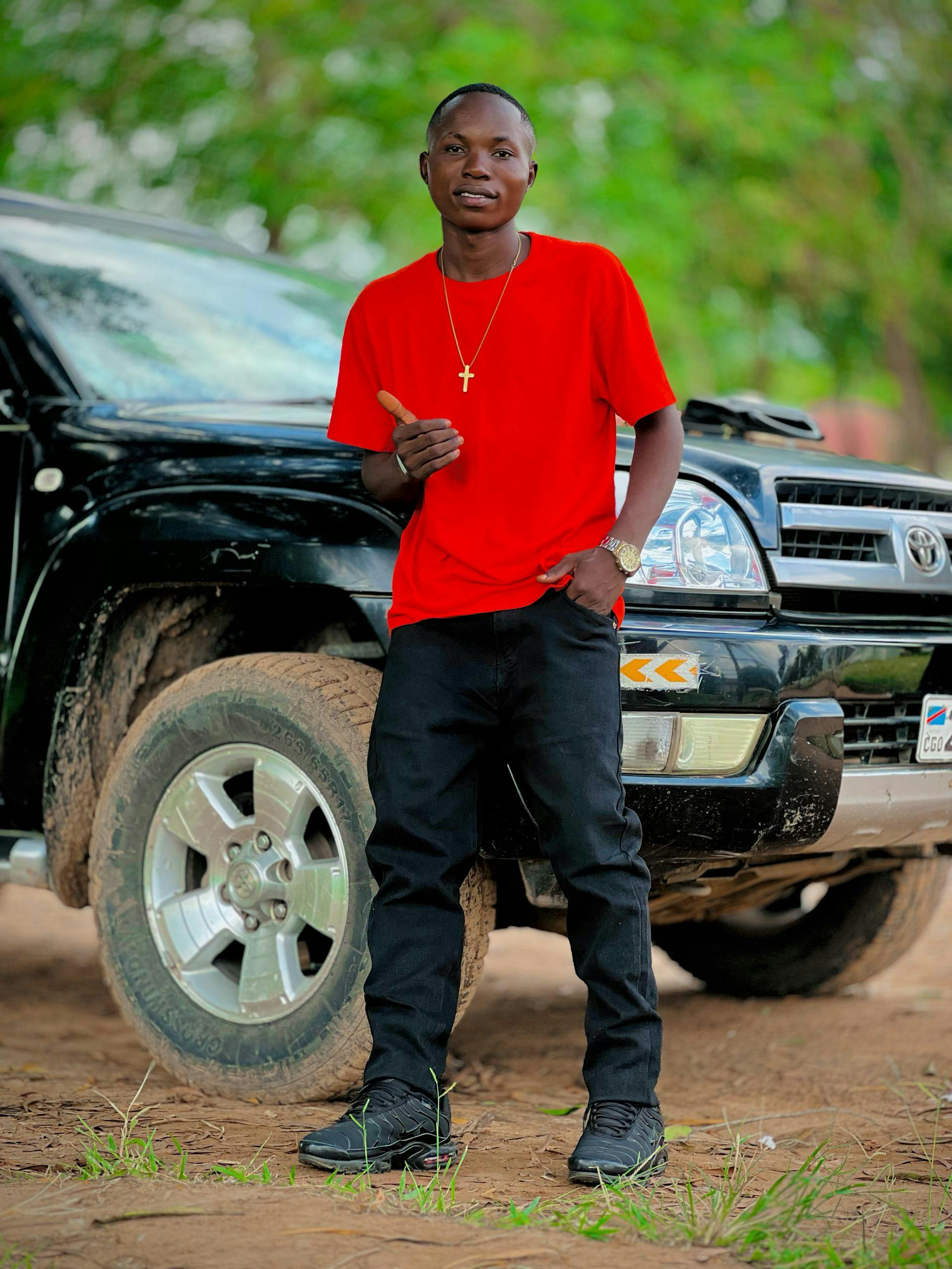 Confident young man in red shirt poses next to an SUV amidst greenery.