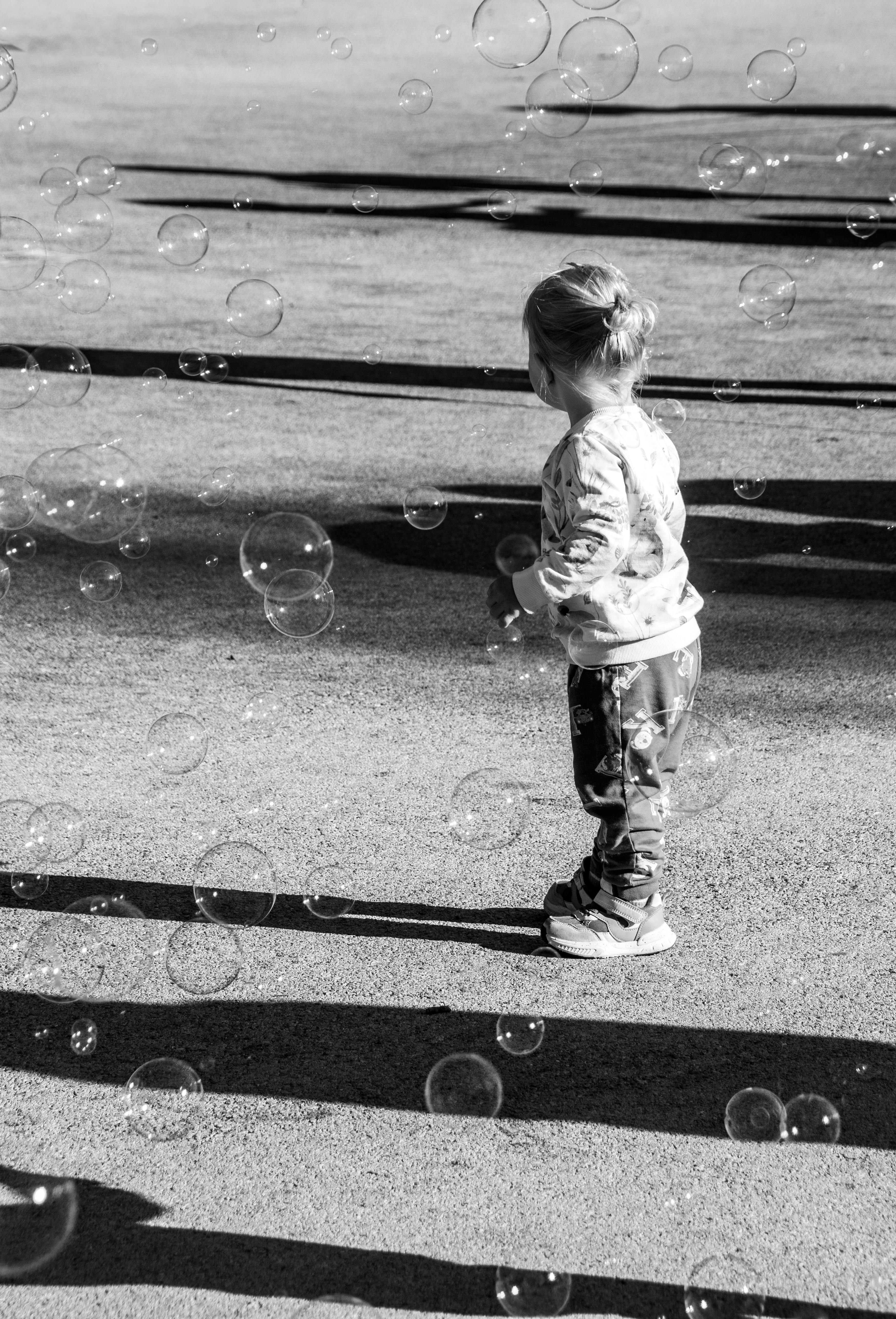 A child stands among bubbles, casting long shadows on the ground in bright sunlight.