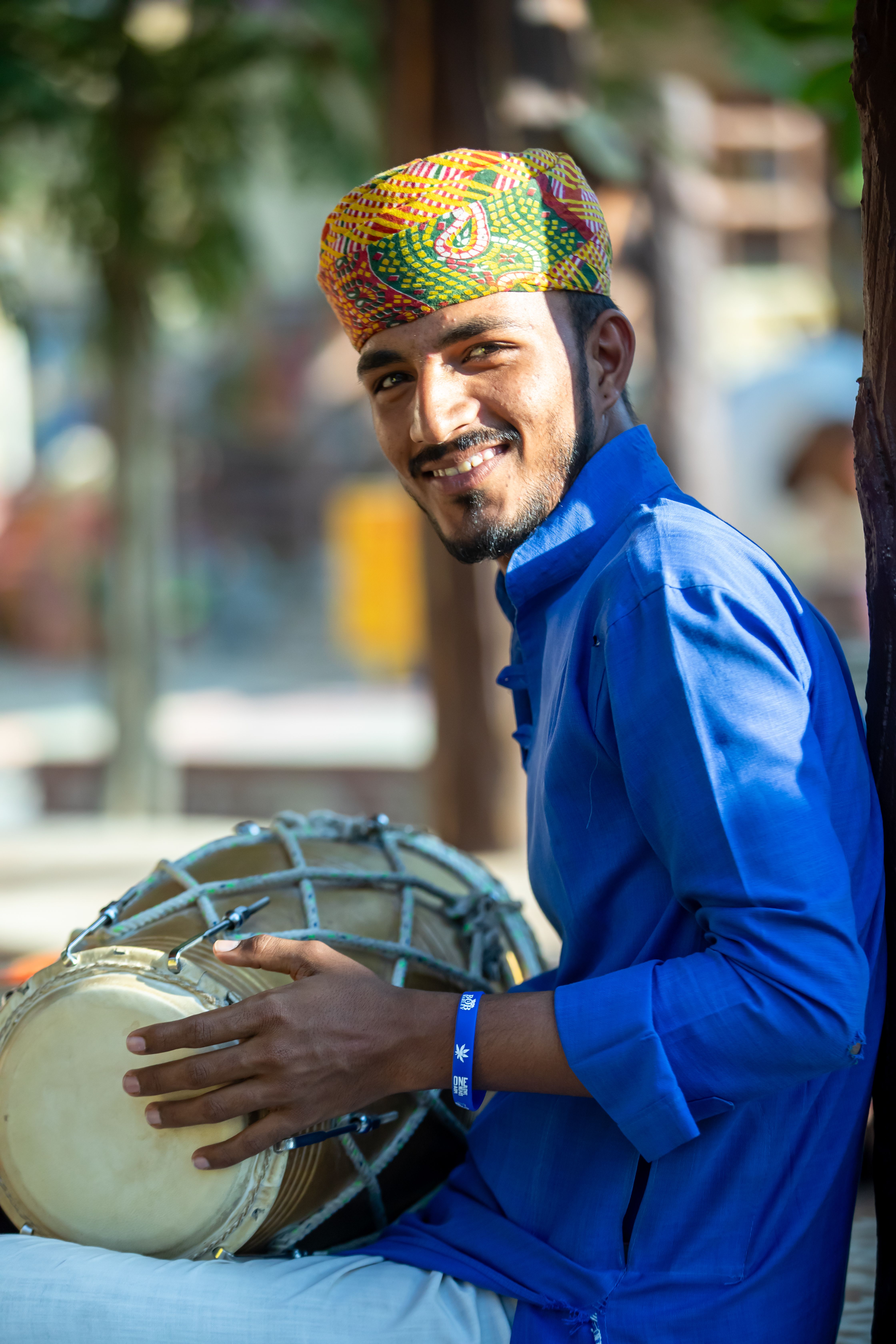 A joyful man wearing traditional Rajasthani clothing and playing a dhol outdoors.