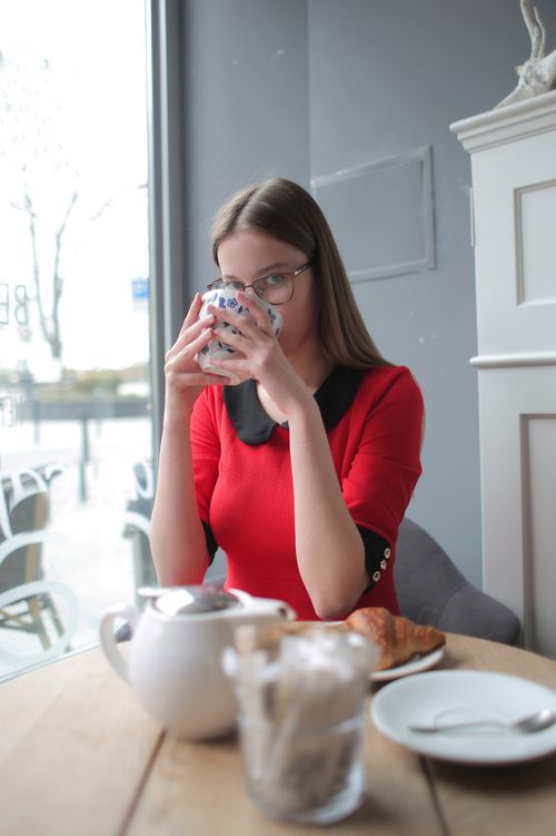 Free A woman in red enjoying coffee at a café table by a window on a calm morning. Stock Photo