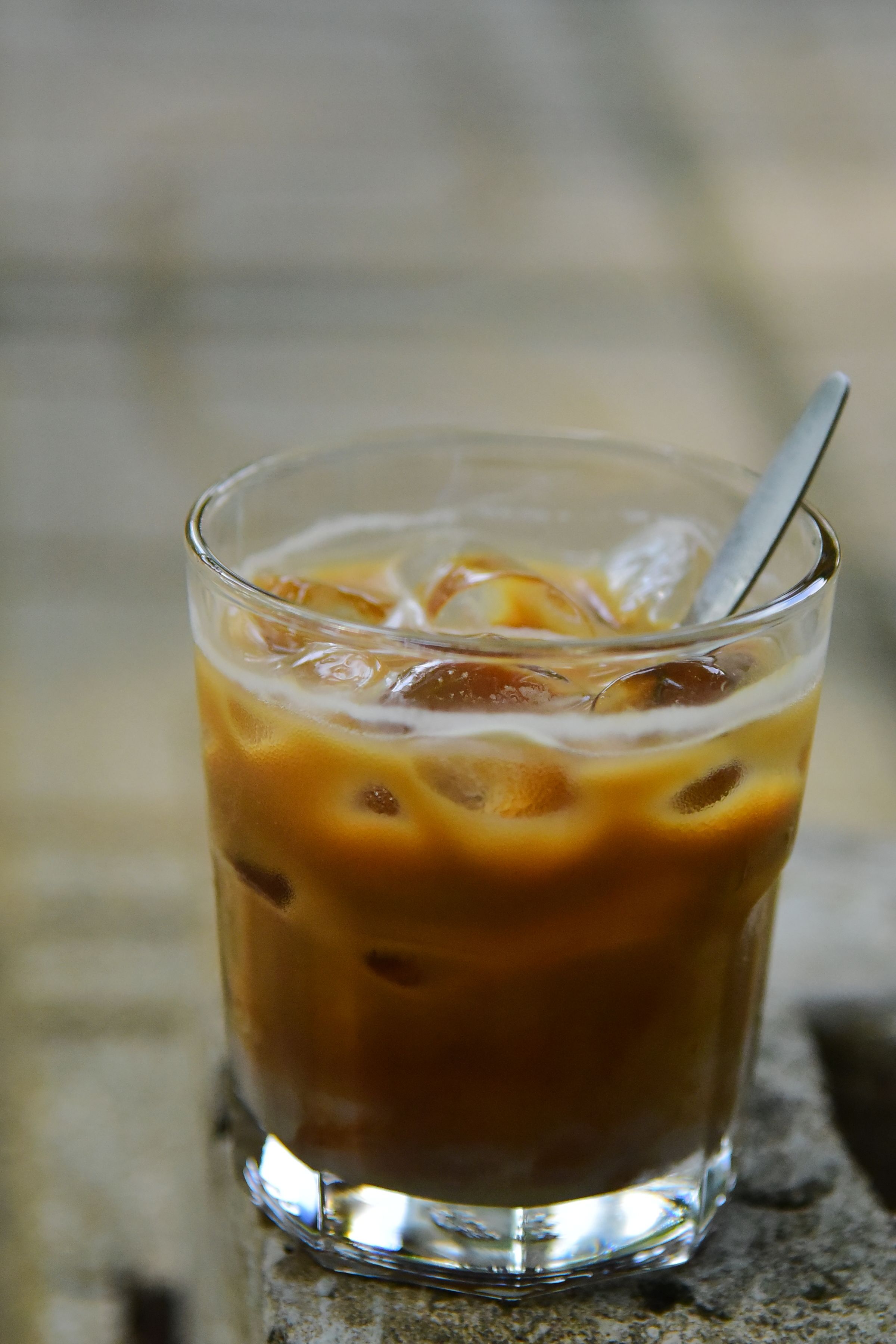 A glass of iced coffee with ice cubes and a spoon outdoors on a sunny day.