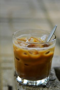 A glass of iced coffee with ice cubes and a spoon outdoors on a sunny day.