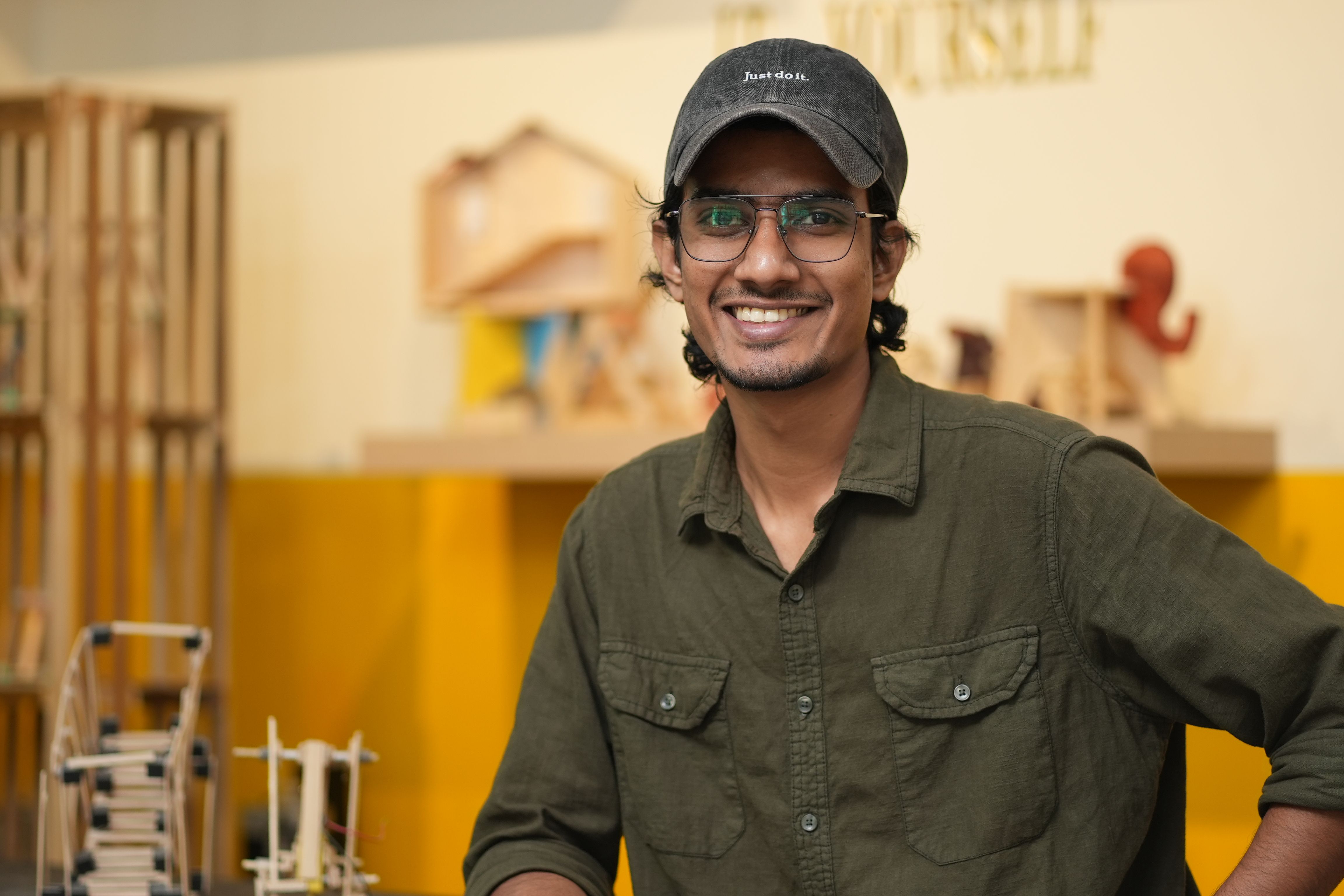 A cheerful young man wearing glasses and a cap relaxes in a creative indoor setting.