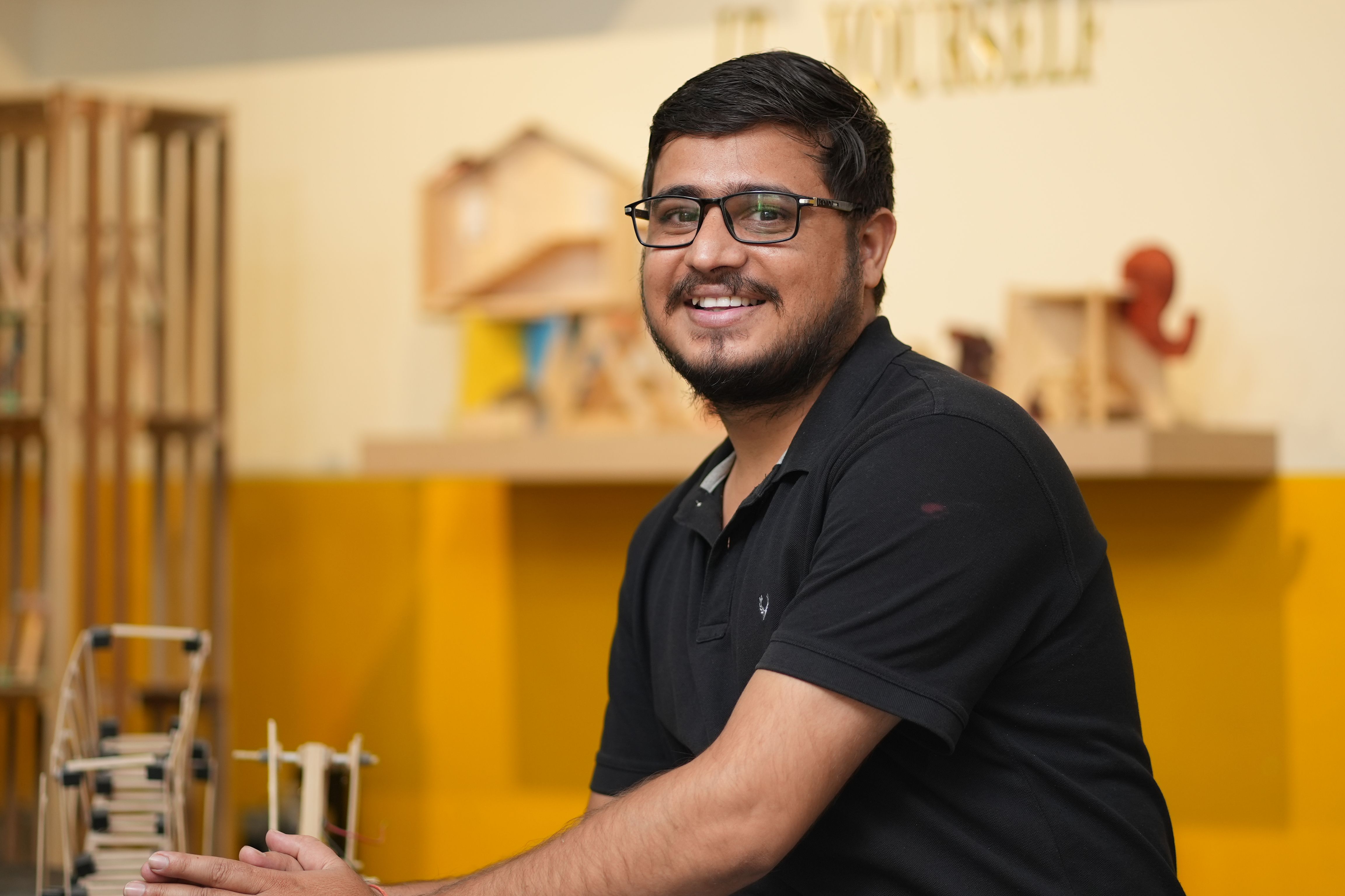 Smiling man in a creative workspace, showcasing DIY models on a wooden shelf.