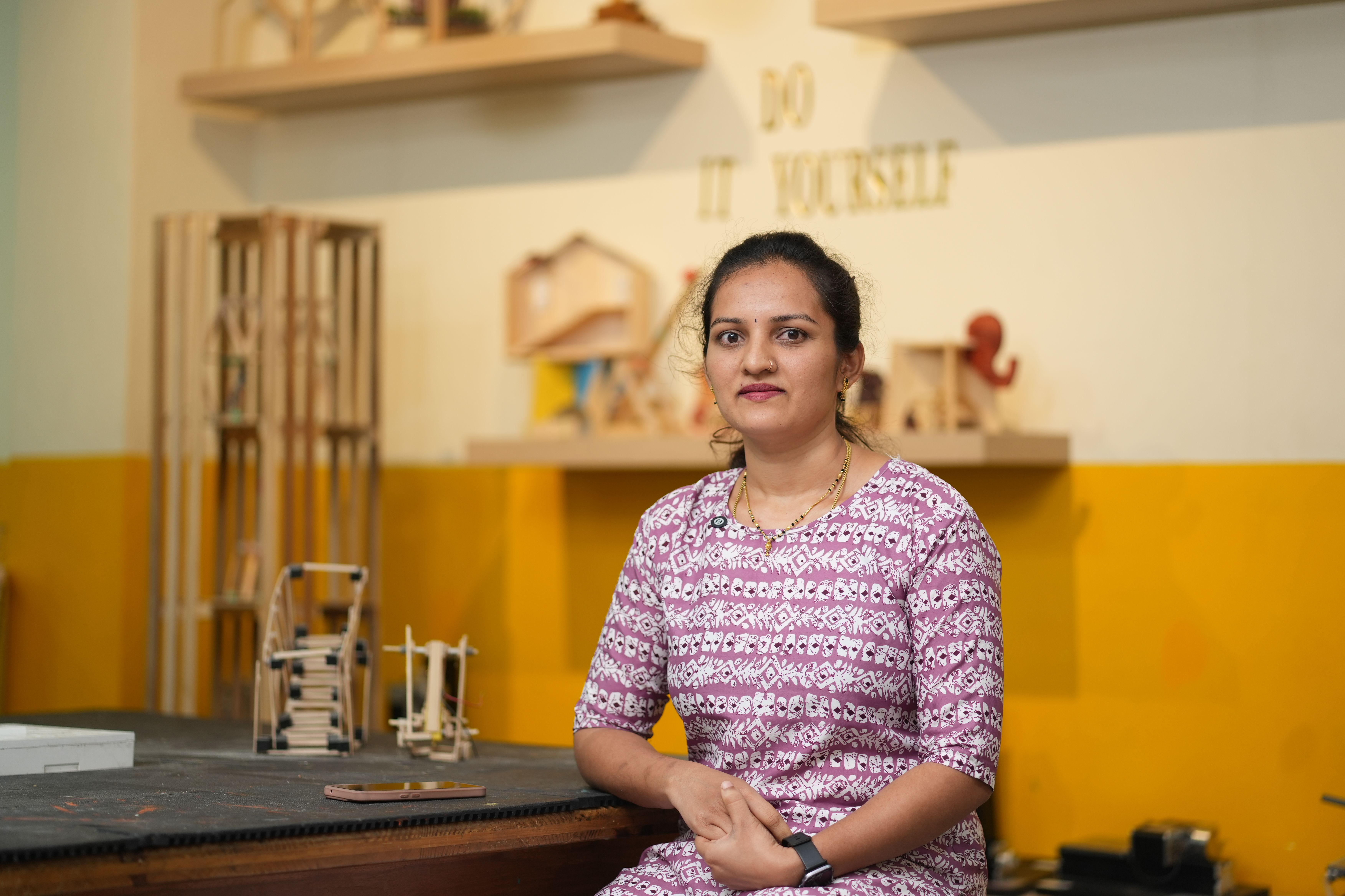 Woman sitting in a DIY workshop surrounded by wooden craft projects.