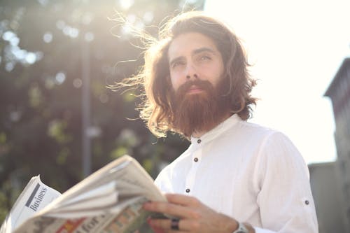 Free Young bearded man enjoying a sunny day while reading a newspaper outdoors. Stock Photo