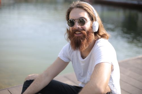 Free A happy man with a beard enjoys music by a serene lake, sitting outdoors. Stock Photo