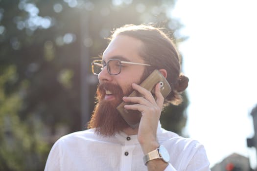 Smiling bearded man with glasses and bun using a smartphone outdoors on a sunny day.
