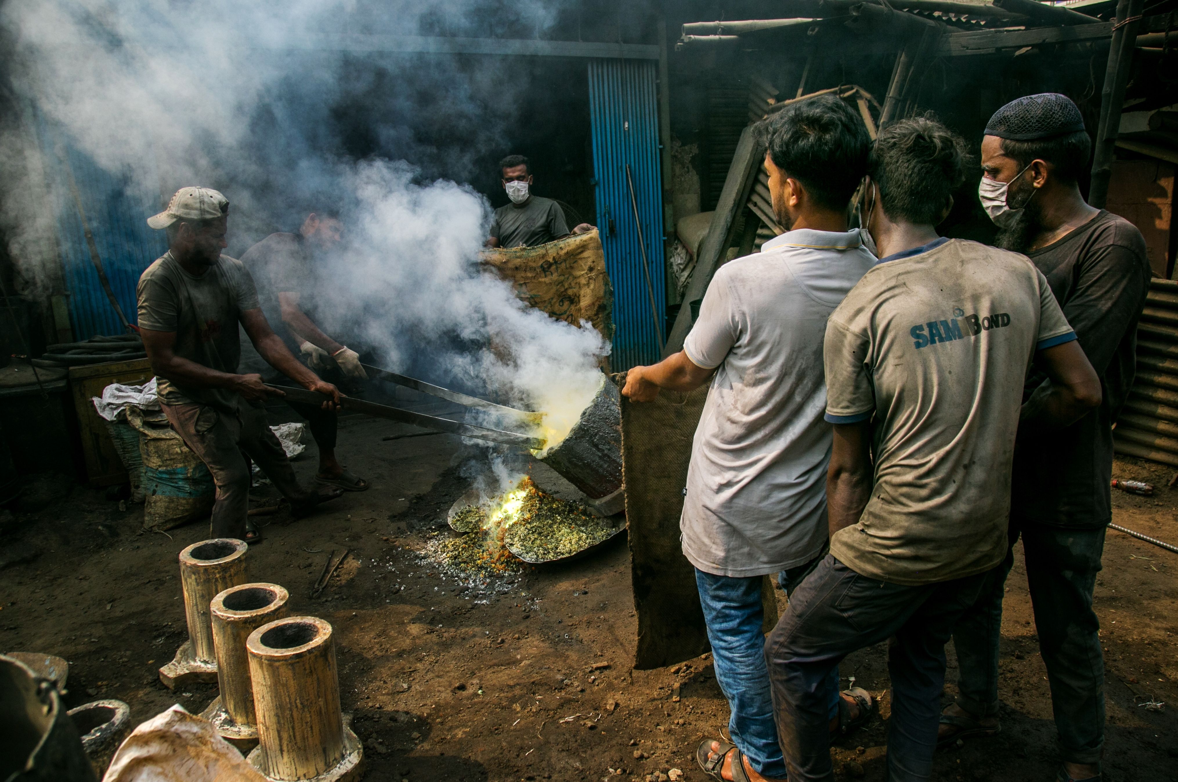 A group of men work together in a smoky metal casting workshop, handling molten metal.