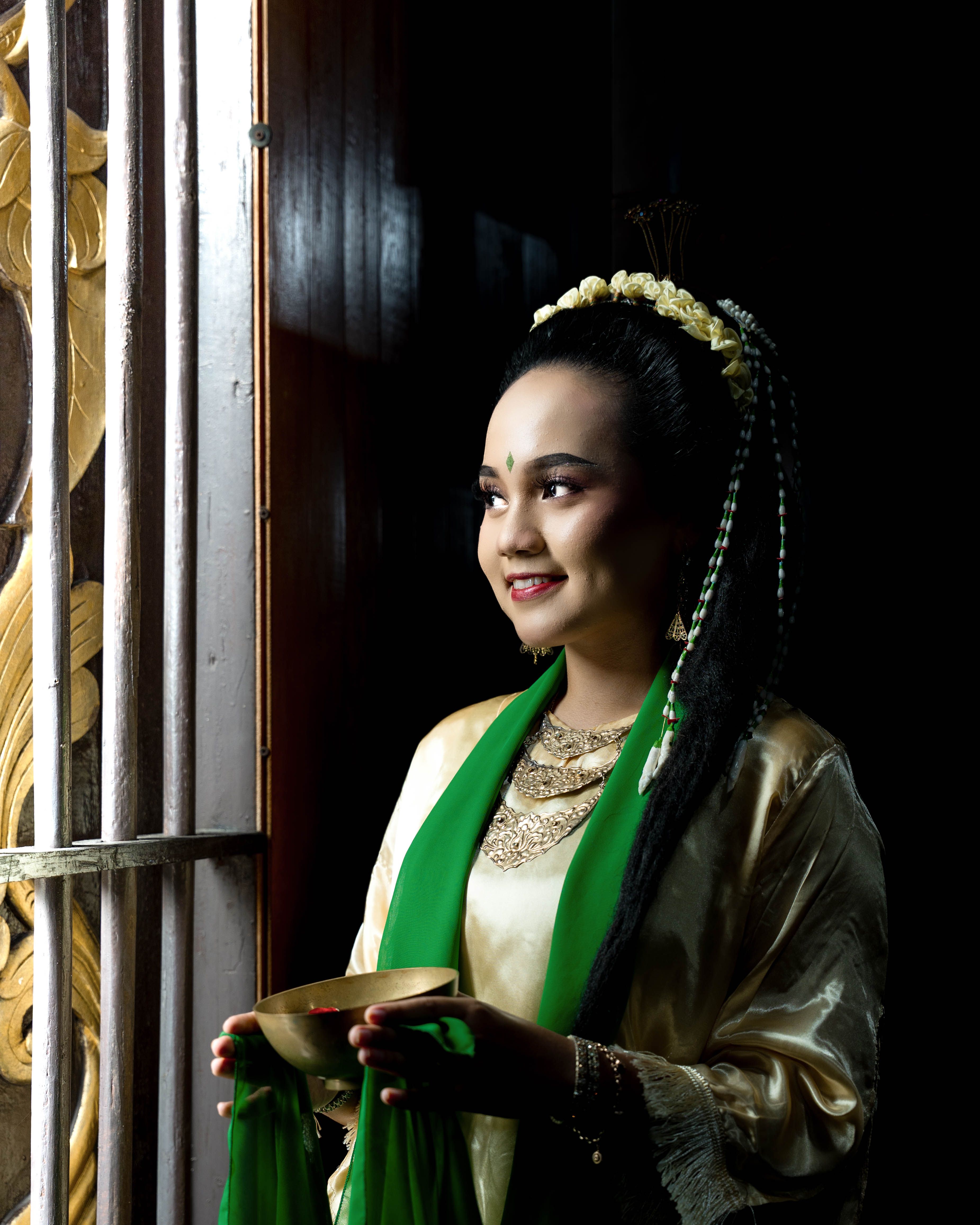 A woman in traditional Indonesian attire holding a tray, standing by an ornate door with soft lighting.